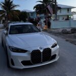 Weißer BMW vor tropischer Villa mit Palmen und Meerblick bei Dämmerung.