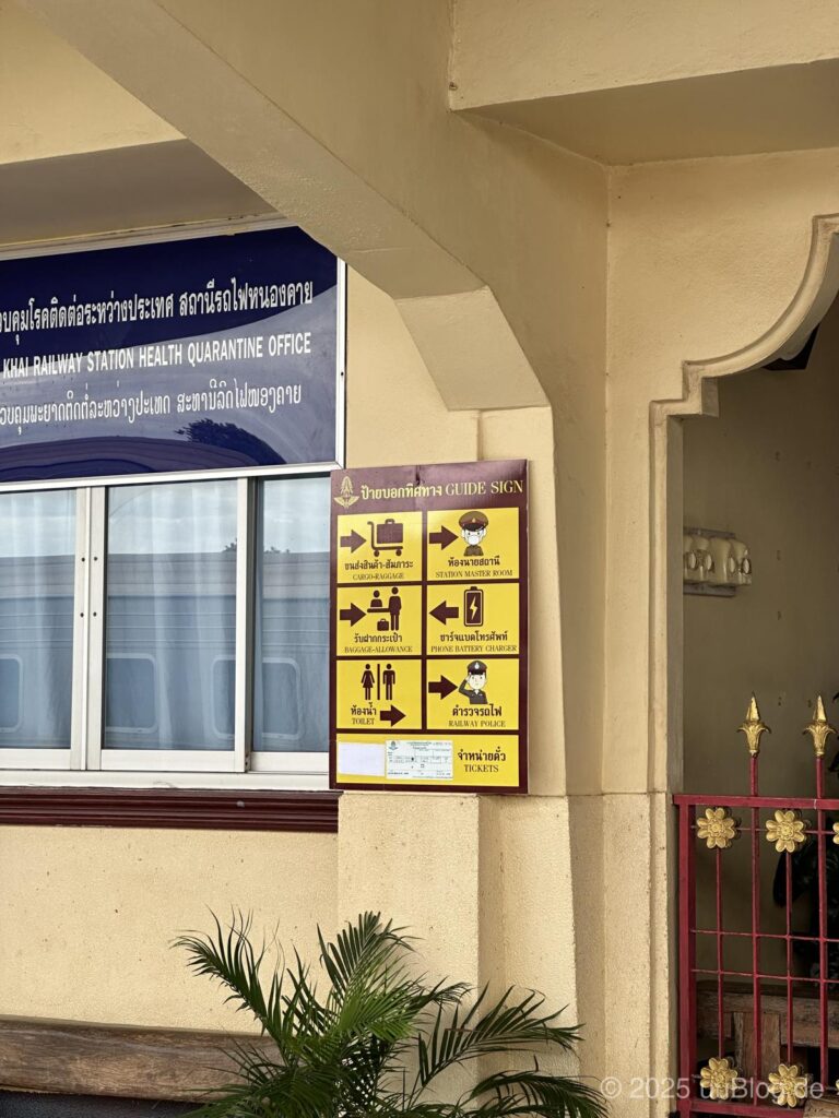 Gelbes Hinweisschild am Nong Khai Bahnhof mit verschiedenen Piktogrammen und thailändischer Beschriftung.