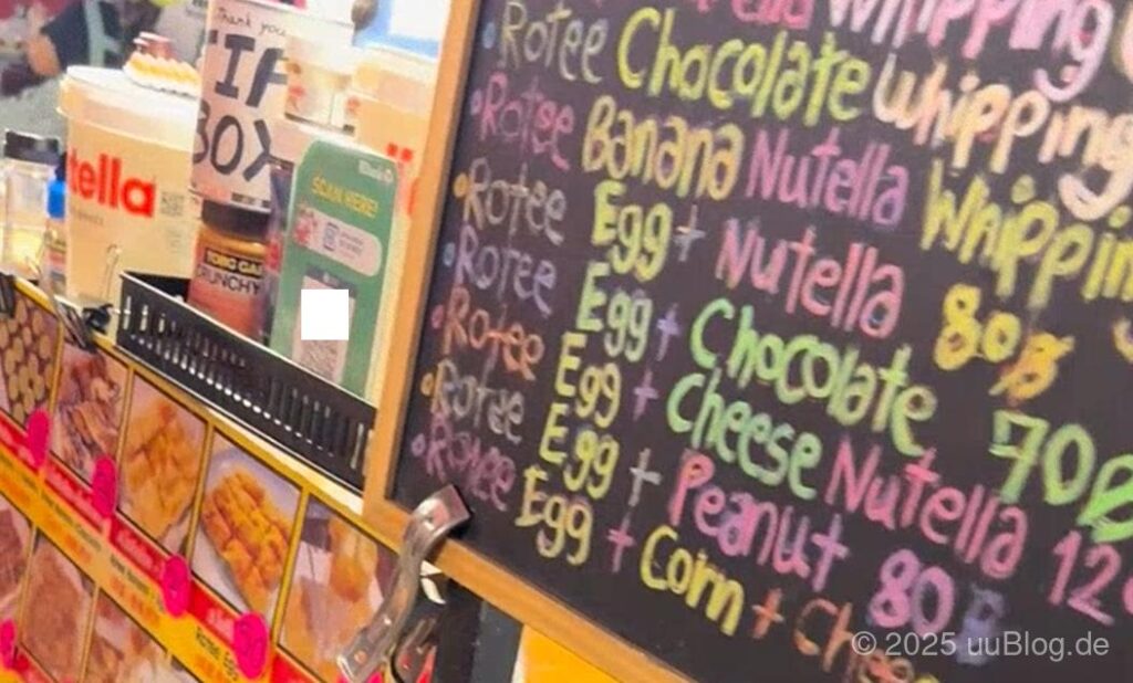 Eine bunte Tafel mit verschiedenen Roti-Menüs und Preisen, daneben Nutella-Gläser und QR-Code-Box für Zahlungen.