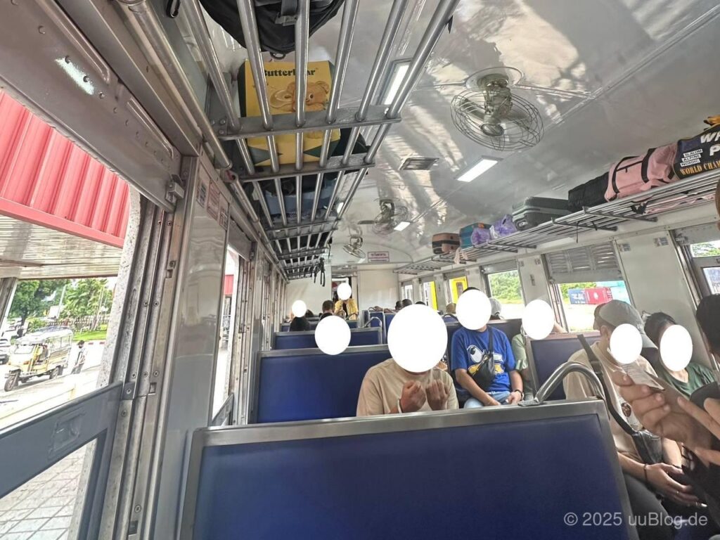 Innenraum eines thailändischen Zugwagons mit Passagieren auf blauen Sitzen und Gepäckfächern oben.