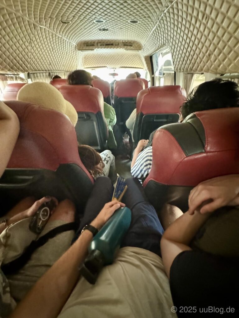 Ein enger Bus mit roten Sitzen, viele Menschen sitzen dicht aneinander und einige schlafen oder ruhen.