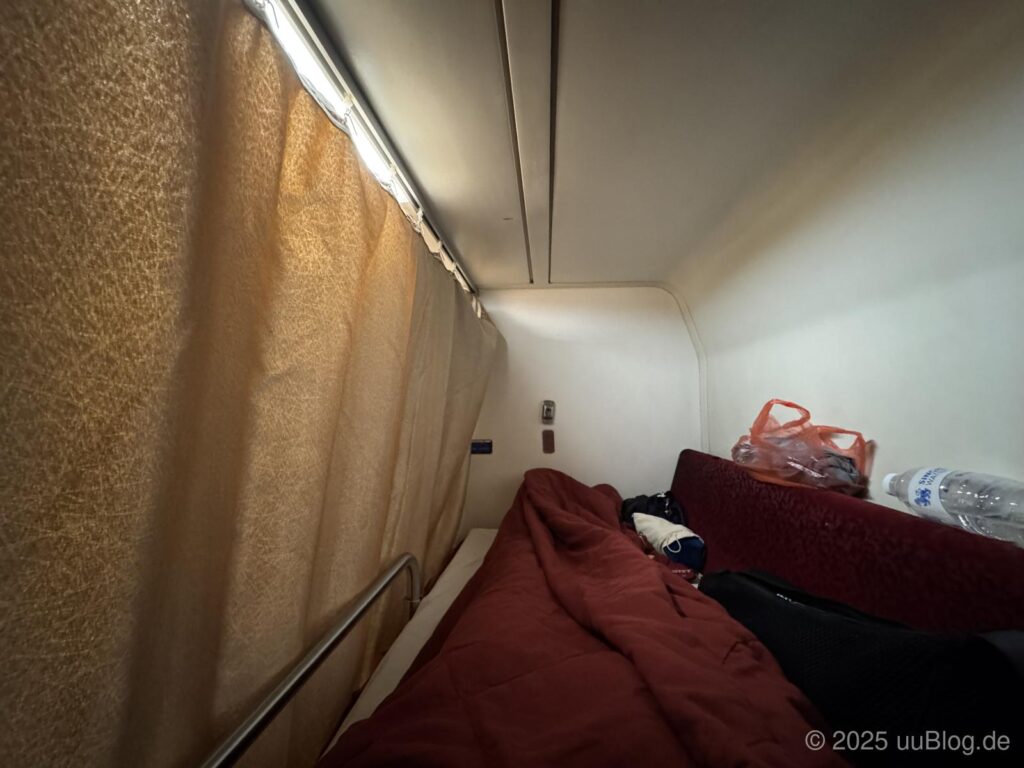 Ein Zugabteil mit Vorhang, Bett und Gepäck.