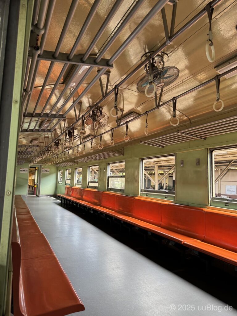 Ein leerer Zugwagen mit roten Sitzbänken, hellgrünen Wänden und Lüftern an der Decke.