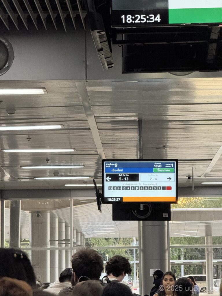 Menschenmenge in einem Bahnhof mit einer Anzeigetafel für einen Zug nach Chiang Mai und der Uhrzeit 18:25.